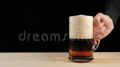 新鲜啤酒。 手从黑色背景的木桌上拿着一杯美味的工艺啤酒在啤酒杯里。 冷鲜啤酒